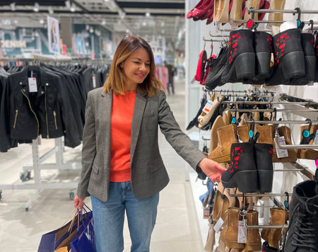 Adult beautiful happy woman standing in store looking at shoes. Smiling Caucasian pretty female buyer buy boots in shopping mall. Clothes, fashion shop. Season sale conceptの写真素材