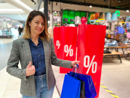 One Caucasian happy beautiful woman standing in shopping mall with bags in hand looking at camera and showing thumb up. Pretty woman shopaholic near store with salesの写真素材