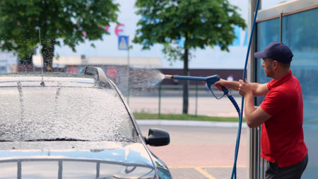 Portrait of handsome concentrated man in cap and red t-shirt washing his car outdoors using high pressure water. Guy cleaning automobile with foam in a self service car wash. Car wash stationの写真素材