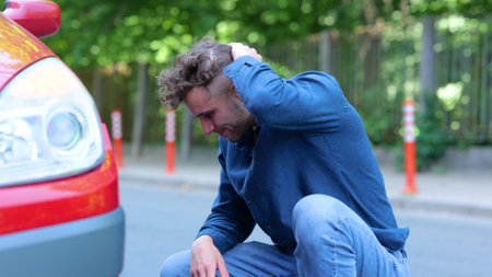 Side view of young male standing near his car looking at tire feeling sad. Close up. Man driver looks at flat tire in red auto. Damaged vehicle. A breakdown on road trip. Car problem.の写真素材