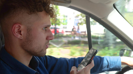 Young handsome serious man driving a car and typing on smartphone while looking at road. Side view....