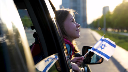 Joyful little girl holding Israeli flag in hand looking out of car window. Close up shot of small pretty child travels in Israel. Patriotic. Family trip. Travel concept. Slow motionの写真素材