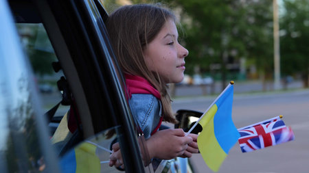 Caucasian kid teen looks out car window in front seat with Ukrainian and British flags in hand....