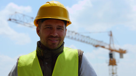Close up of handsome cheerful man builder in yellow hard hat looking at camera and smiling outdoors....