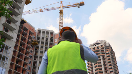 Back view of man builder in uniform standing in front of construction site looking at building...