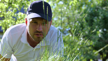 Handsome young Caucasian man cutting green grass with scissors in private yard. Close up of...