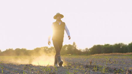 Joyful handsome male farmer walks in rubber boots on land through small green sprouts of wheat. Man cultivator walking on dry soil at own farm on sunset. Farmland, agronomist conceptの写真素材
