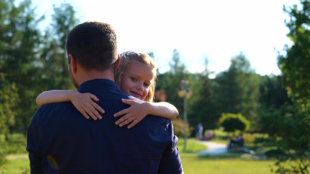 Back view of man walking in nature holding his little daughter in arms. Pretty child girl smiling to camera hugging father outdoor in park. Summer weekend. Family day. Childhood conceptの写真素材