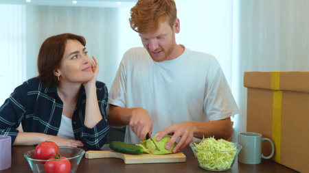 Happy Caucasian couple standing in new house preparing food together. Cheerful couple man and woman in new own house. Husband cutting vegetables and eating cabbage in kitchen. Slow motionの写真素材