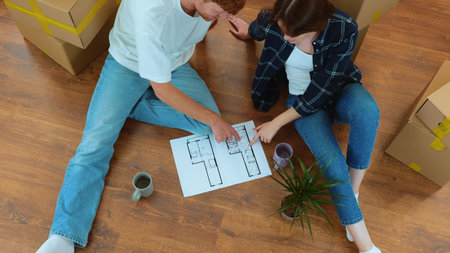 Cheerful man and woman sitting on the floor in new house with coffee and home project discussing...