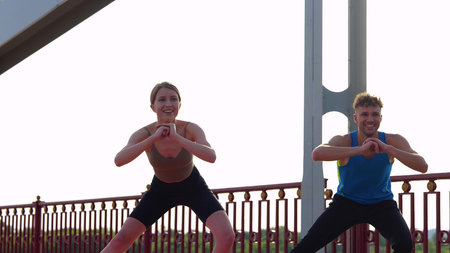 Cheerful man and woman in sportswear working out together outdoors doing side lunges. Caucasian...