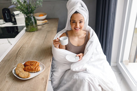 Happy young woman in white towel on the head holding cup of coffee and sitting on the kitchen at home. Morning routine. Lifestyle conceptの写真素材