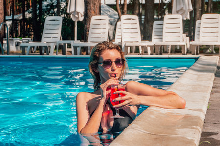 Happy woman in sunglasses drinking the cocktail while standing in the swimming pool outdoors. Copy space. Summer, vacation and lifestyle conceptの写真素材