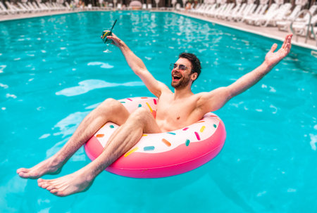 Happy young man holding hands to the side while resting in the swimming pool with cocktail....