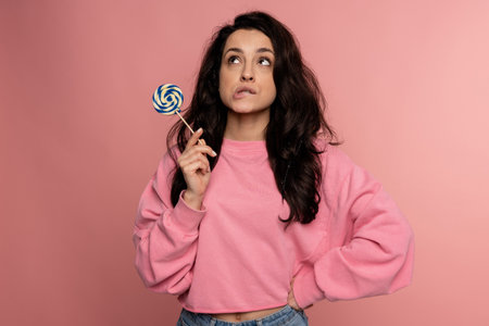 Cute pensive young woman with a sugar candy staring upward while standing in front of the camera on the pink background. Sweet food conceptの写真素材