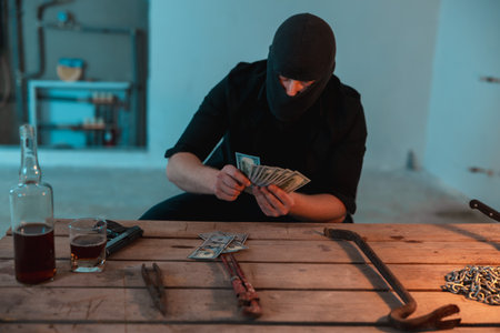 Robber in a black balaclava counting money in the basement. Gun and alcohol on the table. Crime, kidnapping, violence conceptの写真素材