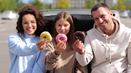 Camera moving back from happy Caucasian lovely family parents and teenager daughter smiling to camera holding in hands donuts. Couple with teen kid girl sitting outdoor in car trunkの写真素材