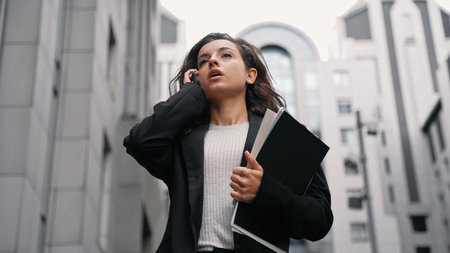 Young Caucasian businesswoman talking by phone, carrying laptop, and files in her hand, walking...