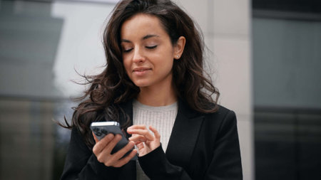 Caucasian businesswoman standing next to office buildings, smiling, looking around, teaching her dark long hair, scrolling her smartphone.の写真素材