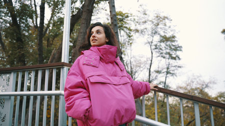 Pretty young woman going down stairs while walking in the park, holding on to the railing, wearing stylish pink jacket. Seasonal style, autumn season concept. Slow motionの写真素材