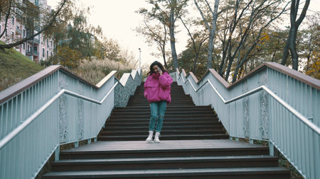 Smiling pretty woman in stylish jacket talking on the smartphone while walking down the stairs in the park. Seasonal style, autumn season concept. Slow motionの写真素材