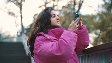 Pretty woman wearing fashionable jacket while walking in the city park and making photo on the smartphone. Seasonal style, autumn season concept. Slow motionの写真素材