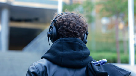 Young man in black wear with backpack and wireless headphones listening to music while walking in...