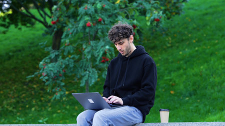 Young man drinking coffee and typing on the laptop, sitting in the city park. Autumn season. Education, working concept. Slow motionの写真素材