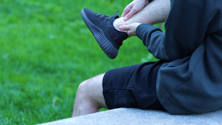 Side view young Caucasian man sitting on concrete bench and his leg hurt after workout. Outdoor....