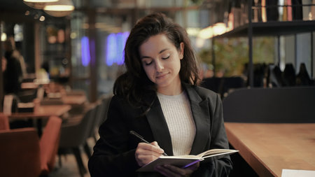 Young Caucasian female with dark long hair and beautiful smile sitting in cafe and drawing her...