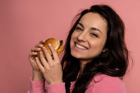 Pleased brunette with a delicious cheeseburger in her hands smiling during the studio photo shoot....