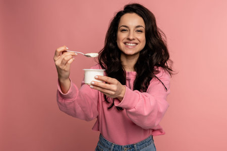 Smiling pleased dark-haired young lady eating an appetizing dairy product in front of the camera on...