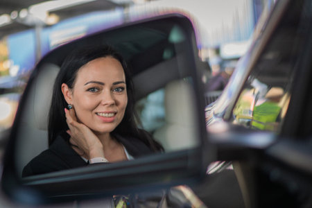 Happy beautiful young woman looking in the side view mirror while sitting in her car. People,...