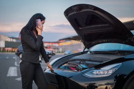 Young woman opening car hood and calling to insurance to help after the car breaks down outdoors. Waiting for help. Car service, transportation conceptの写真素材
