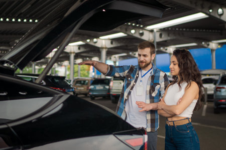 Smiling male instructor and young lady standing near car with open trunk on the parking. Driving test, driver courses, exam conceptの写真素材