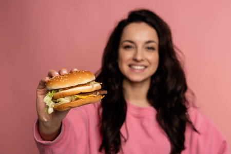 Pleased young dark-haired pretty lady posing with her favorite sandwich during the studio photo shoot. Fast food consumption conceptの写真素材