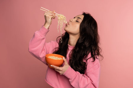 Hungry dark-haired young female snacking on tasty pasta with her eyes closed during the photo shoot on the pink background. Japanese cuisine conceptの写真素材