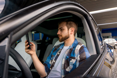 Smiling bearded young man sitting in his car while holding smartphone and reading message. Travel,...