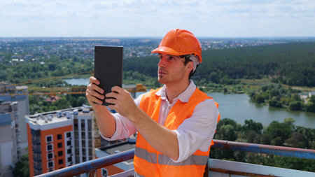 Young male builder standing on the roof while making video of construction site with digital tablet....