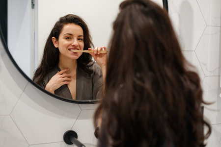 Beauty young woman looking in the big mirror at her bathroom while brushing her teeth. Focus on the reflection in the mirror. Routine, self-care, lifestyle conceptの写真素材