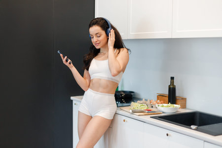 Pretty young woman in underwear enjoying music on headphones while standing in the kitchen of her apartment. Lady posing near the table with food. Lifestyle conceptの写真素材