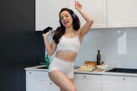 Smiling young female listening to music at home while standing on the kitchen and holding mobile phone. Lifestyle conceptの写真素材