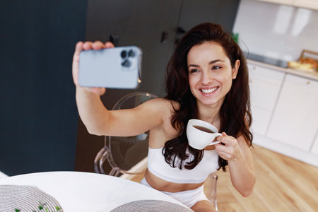 Happy young woman sitting at home, wearing underwear while using phone and taking photo, enjoying tea in the morning. Lifestyle, morning routine conceptの写真素材