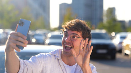 Happy young man holding smartphone, having video call, waving hand and showing V sign, car on the background. Transport, technology and lifestyle concept. Slow motionの写真素材