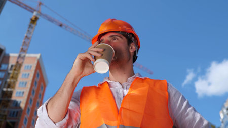 Builder in working uniform drinking coffee while standing construction site. Business, building, industry concept. Slow motionの写真素材