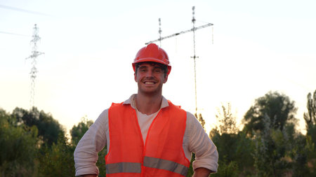Happy young male engineer standing outdoors in front of a high voltage power lines. Distribution and...