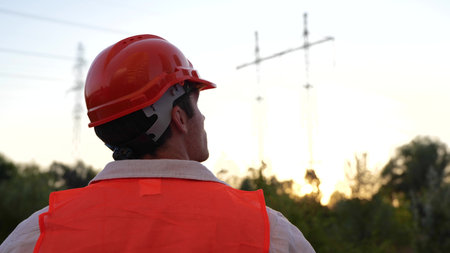 Young engineer standing outdoors while looking at the high voltage power lines. Back view. Energy...