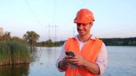 Happy male engineer in safety helmet typing on the smartphone. Highest voltage transmission. River on the background. Energy industry concept. Slow motionの写真素材
