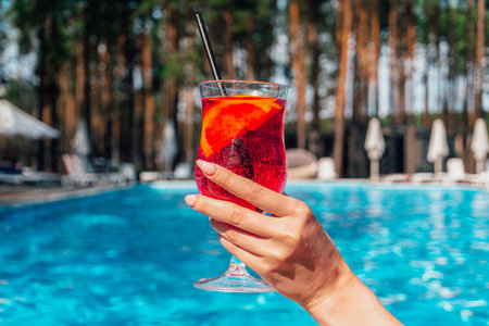 Female hand holding cocktail. Swimming pool on the background. Cropped, copy space. Summer, party, vacation and lifestyle conceptの写真素材