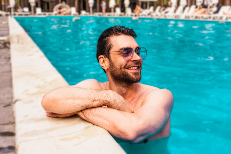 Smiling muscular young man in sunglasses looking away while enjoying rest in the swimming pool outdoors. Summertime, holidays, lifestyle conceptの写真素材